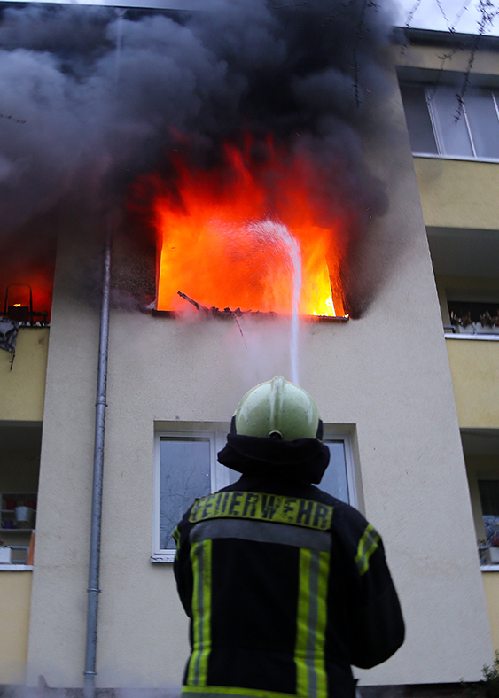 Symbolbild für Notfallnummern - Flammen schlagen aus der Wohnung eines Mehrfamilienhauses. Ein Feuerwehrmann löscht. Foto: Stefan Rampfel (rammi76 Adobe Stock)